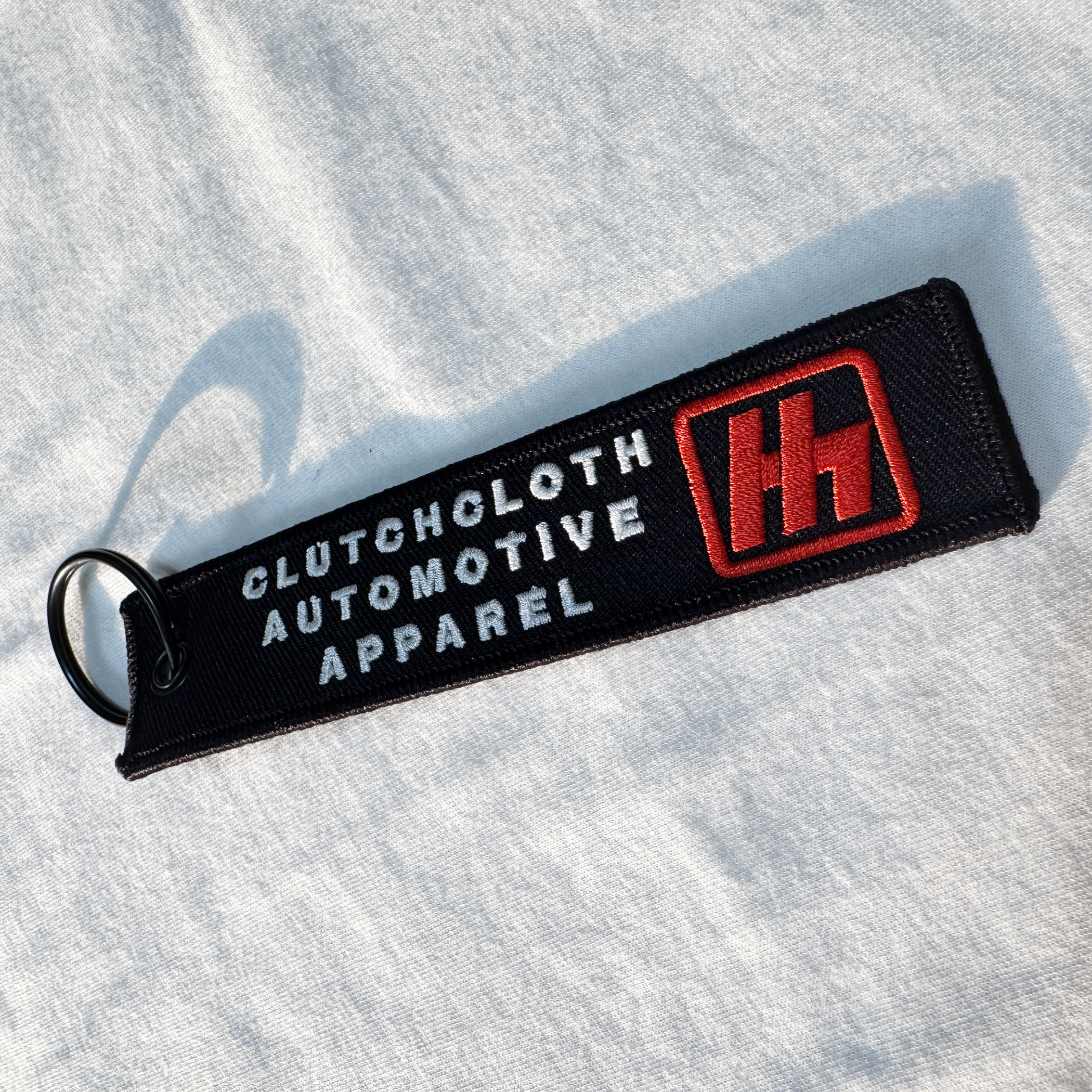 Black keychain with 'Clutcholoth Automotive Apparel' text and logo on a white fabric background