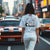 Woman wearing Cobro x Clutch Automotive Graphic T-Shirt – Blue tee featuring a detailed white Mustang Cobra design with graffiti-style background and "Cobro" text. Perfect gift for Mustang fans and car enthusiasts.